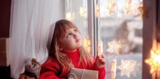 A child in a red sweater holding a gift while looking out a window decorated with lights