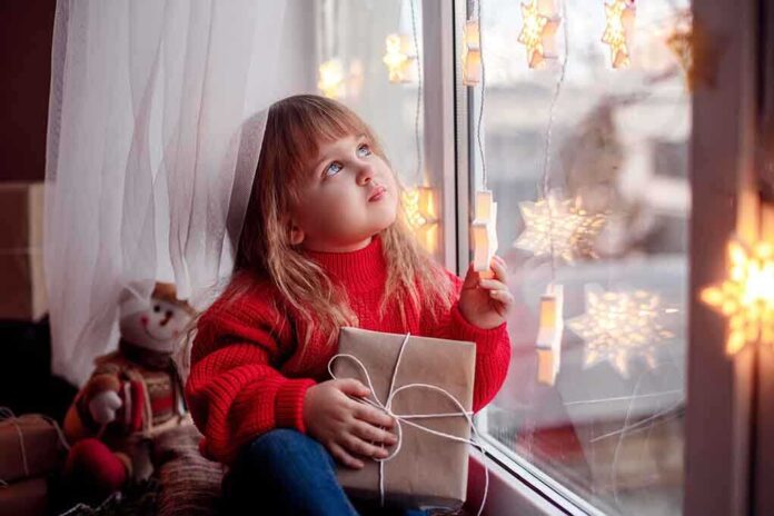 A child in a red sweater holding a gift while looking out a window decorated with lights