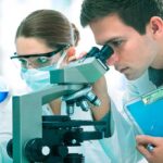 Two scientists conducting an experiment in a laboratory one looking through a microscope and the other holding a flask with blue liquid