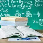 Open textbook on a desk with stacked books and a chalkboard filled with math equations in the background