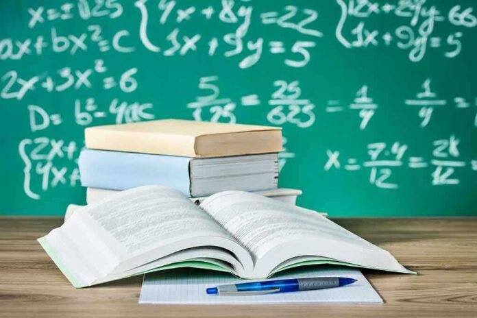 Open textbook on a desk with stacked books and a chalkboard filled with math equations in the background