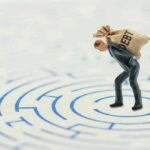 Debt-Haters Caught in Credit Card Frenzy A miniature businessman carrying a sack labeled 'DEBT' in a maze