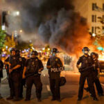 Police officers in riot gear near burning car.