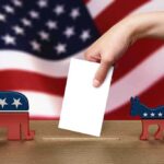 A hand placing a ballot into a box with political symbols on either side against a backdrop of the American flag