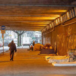 ER Visits TRIPLE: New Drug Nightmare Unleashed People walk under a bridge with homeless encampment.
