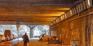 People walk under a bridge with homeless encampment.
