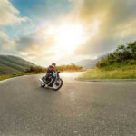Person rides motorcycle on curved mountain road at sunset.