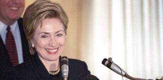 A smiling woman at a podium with microphones, speaking to an audience