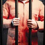 Actor’s Violent Rampage Rocks Mardi Gras Celebration Person in an orange jumpsuit gripping prison bars in a dark cell