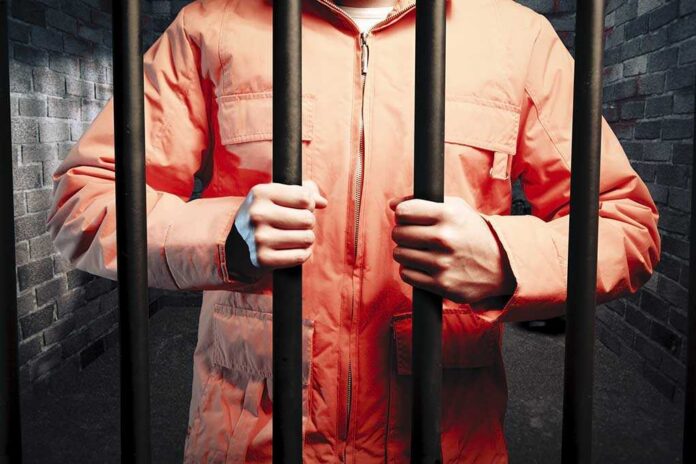 shutterstock_62493325.jpg Person in an orange jumpsuit gripping prison bars in a dark cell