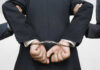 Person handcuffed in suit, escorted by two others.
