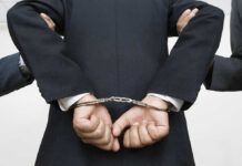 Person handcuffed in suit, escorted by two others.