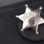 DETECTIVE AMBUSHED — Sheriff’s Brutal Response Stuns Nation A silver sheriff badge resting on a black leather surface
