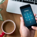 Typing privacy passcode on smartphone near laptop and coffee.