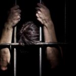 Hands gripping prison bars in a dark setting