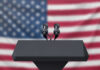 Fraudster Turned TV Star—What’s Fox Planning? Podium with microphones, American flag background.
