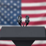 Podium with microphones, American flag background.