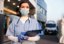 Drug Arsenal Useless Against This Ebola Nurse in scrubs and mask outside hospital holding clipboard.