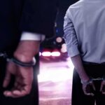 Two men in handcuffs with police lights in the background