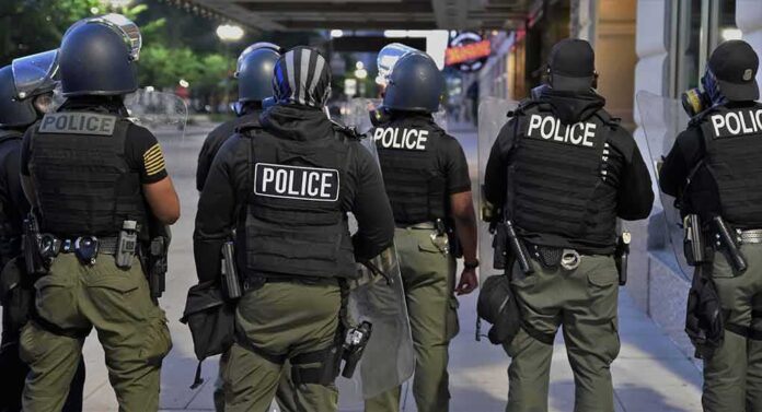 shutterstock_1795453555 Police officers in riot gear stand on a city sidewalk at night
