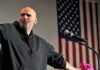Bald man speaking at a microphone with a U.S. flag in the background