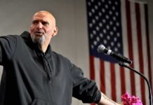 Senator Explodes: ‘Who’s Paying This Outlet?’ Bald man speaking at a microphone with a U.S. flag in the background